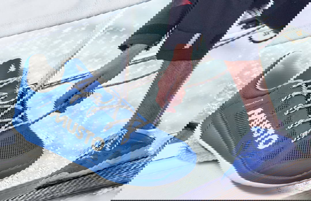 Een blauwe zeilschoen van het merk MUSTO is prominent op de voorgrond te zien, met witte letters en een lichtkleurig vetersysteem. Op de achtergrond knielt een persoon in zeilkleding op een nat bootdek en trekt aan een touw – ook met blauwe zeilc Een blauwe zeilschoen van het merk MUSTO is prominent op de voorgrond te zien, met witte letters en een lichtkleurig vetersysteem. Op de achtergrond knielt een persoon in zeilkleding op een nat bootdek en trekt aan een touw – ook met blauwe zeilc