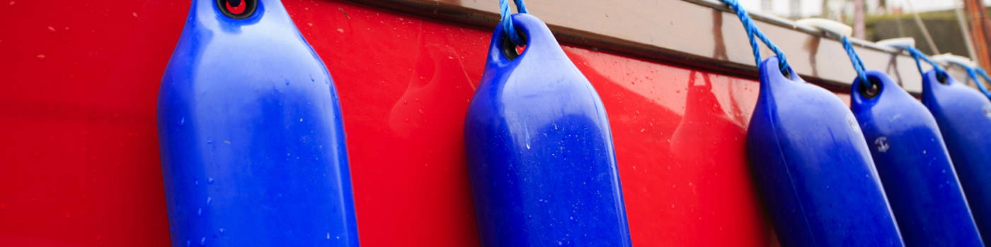 Mehrere blaue zylindrische Bootsfender hängen in gleichmäßigem Abstand entlang der Bordwand eines roten Bootes. Die Fender sind mit blauen Leinen befestigt und schützen das Boot beim Anlegen. Mehrere blaue zylindrische Bootsfender hängen in gleichmäßigem Abstand entlang der Bordwand eines roten Bootes. Die Fender sind mit blauen Leinen befestigt und schützen das Boot beim Anlegen.