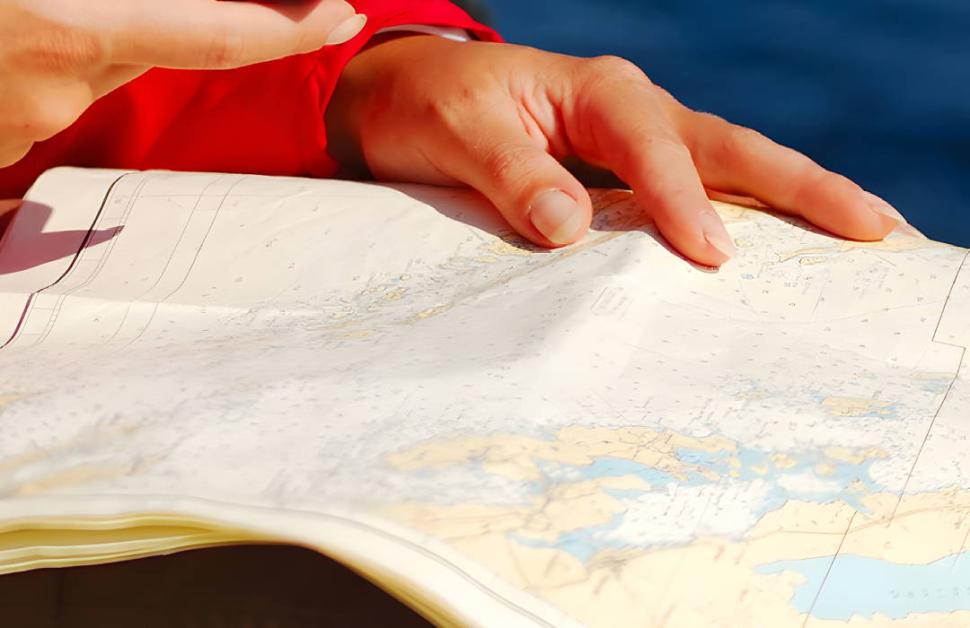 Een hand in een rode zeiljas houdt een uitgespreide zeekaart vast op een boot. De kaart toont gedetailleerde kustlijnen, dieptes en navigatie-informatie. Op de achtergrond is de blauwe zee vaag te zien. Een hand in een rode zeiljas houdt een uitgespreide zeekaart vast op een boot. De kaart toont gedetailleerde kustlijnen, dieptes en navigatie-informatie. Op de achtergrond is de blauwe zee vaag te zien.