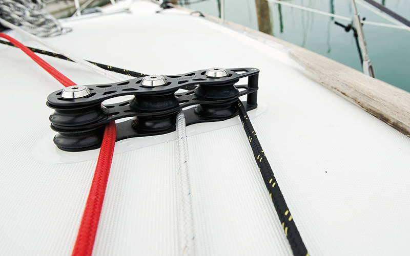 Drie gekleurde zeillijnen (rood, wit, zwart) lopen naast elkaar door een dekdoorvoer met meerdere katrollen op het witte dek van een zeilboot. Drie gekleurde zeillijnen (rood, wit, zwart) lopen naast elkaar door een dekdoorvoer met meerdere katrollen op het witte dek van een zeilboot.