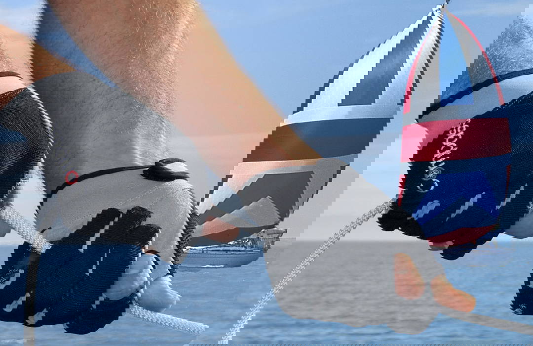 op de achtergrond een boot met een rood-wit-blauwe spinnaker op open zee." "Twee handen met vingerloze Compass-zeilhandschoenen grijpen een touw aan boord van een zeilboot